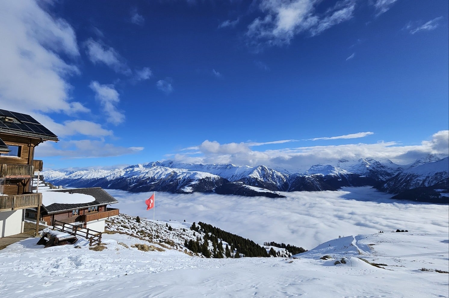 A wooden mountain chalet overlooking snow-covered peaks above a sea of clouds, with a Swiss flag flying in the foreground.