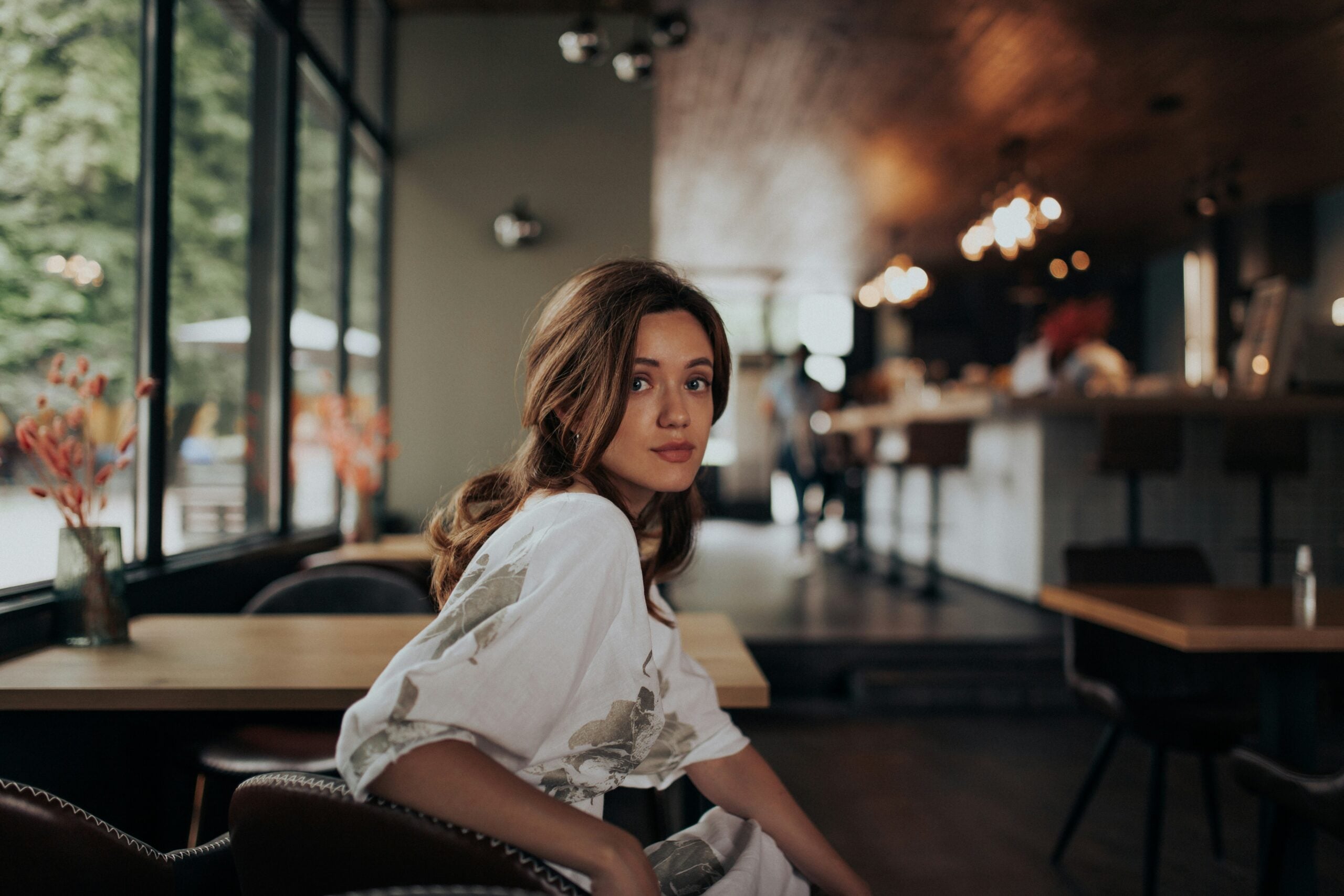 A young woman with brown hair sits in a cozy, modern café, looking over her shoulder toward the camera. The café has wooden tables, soft lighting, and large windows showing greenery outside.