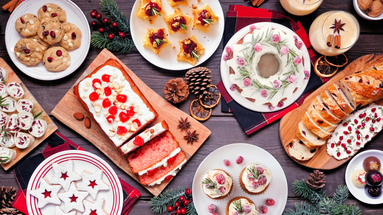 A festive spread of holiday desserts including cookies, frosted loaf cake, a bundt cake, pastries, and eggnog on a wooden table.