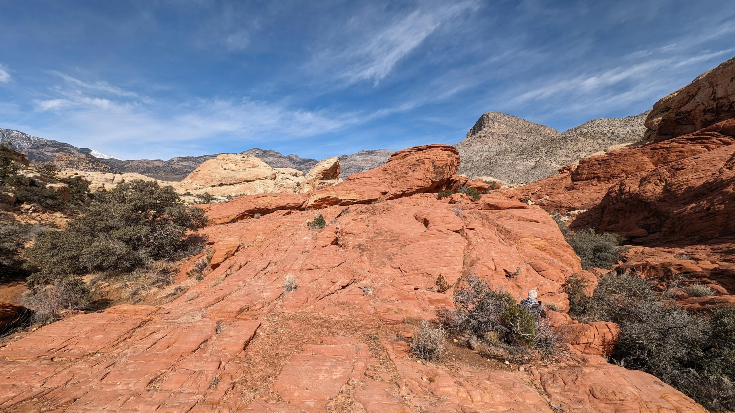 A person sitting on red sandstone rock formations under a bright blue sky with distant mountains.