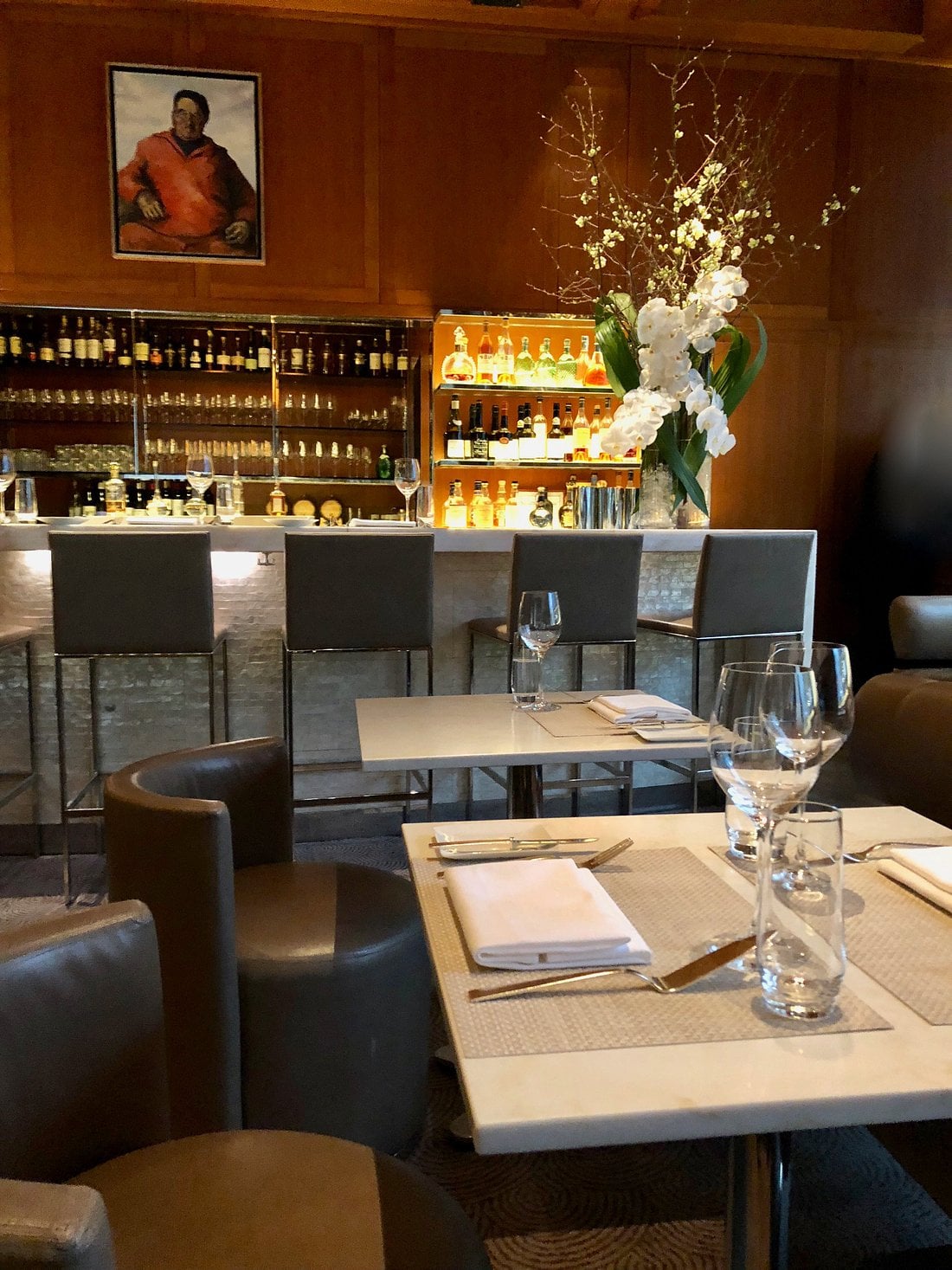 A restaurant interior with bar seating featuring backlit shelves of liquor bottles, white floral arrangements, and tables set with white napkins and glassware.