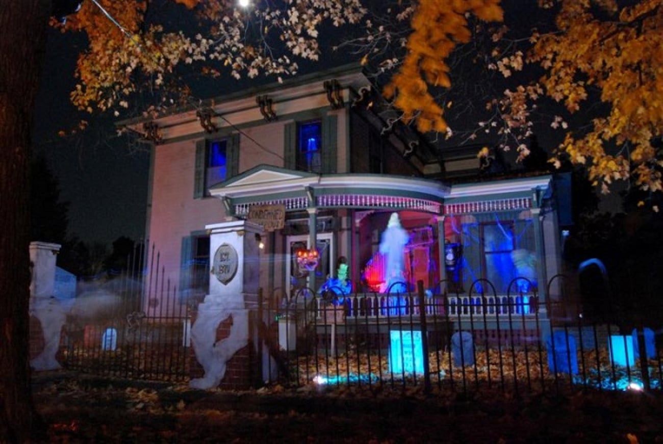 A house decorated for Halloween with a graveyard scene in the front yard, including tombstones, glowing blue lights, and a hanging ghost on the porch.