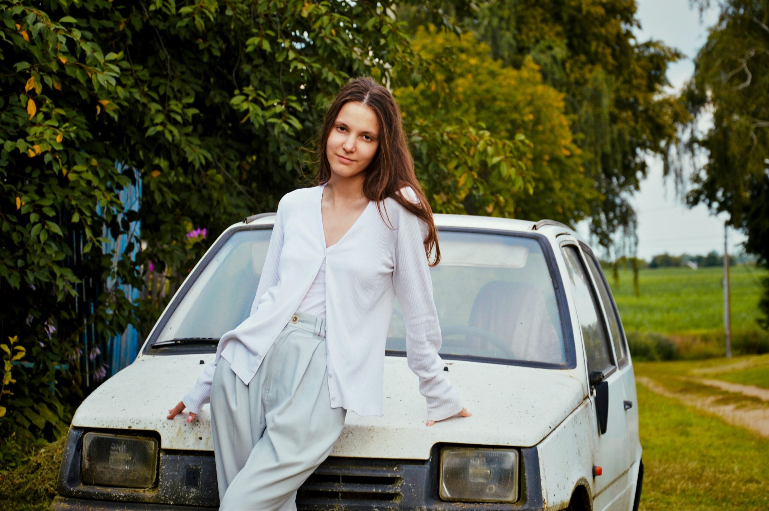 A woman leaning against the hood of a white hatchback car parked in front of trees and a field.