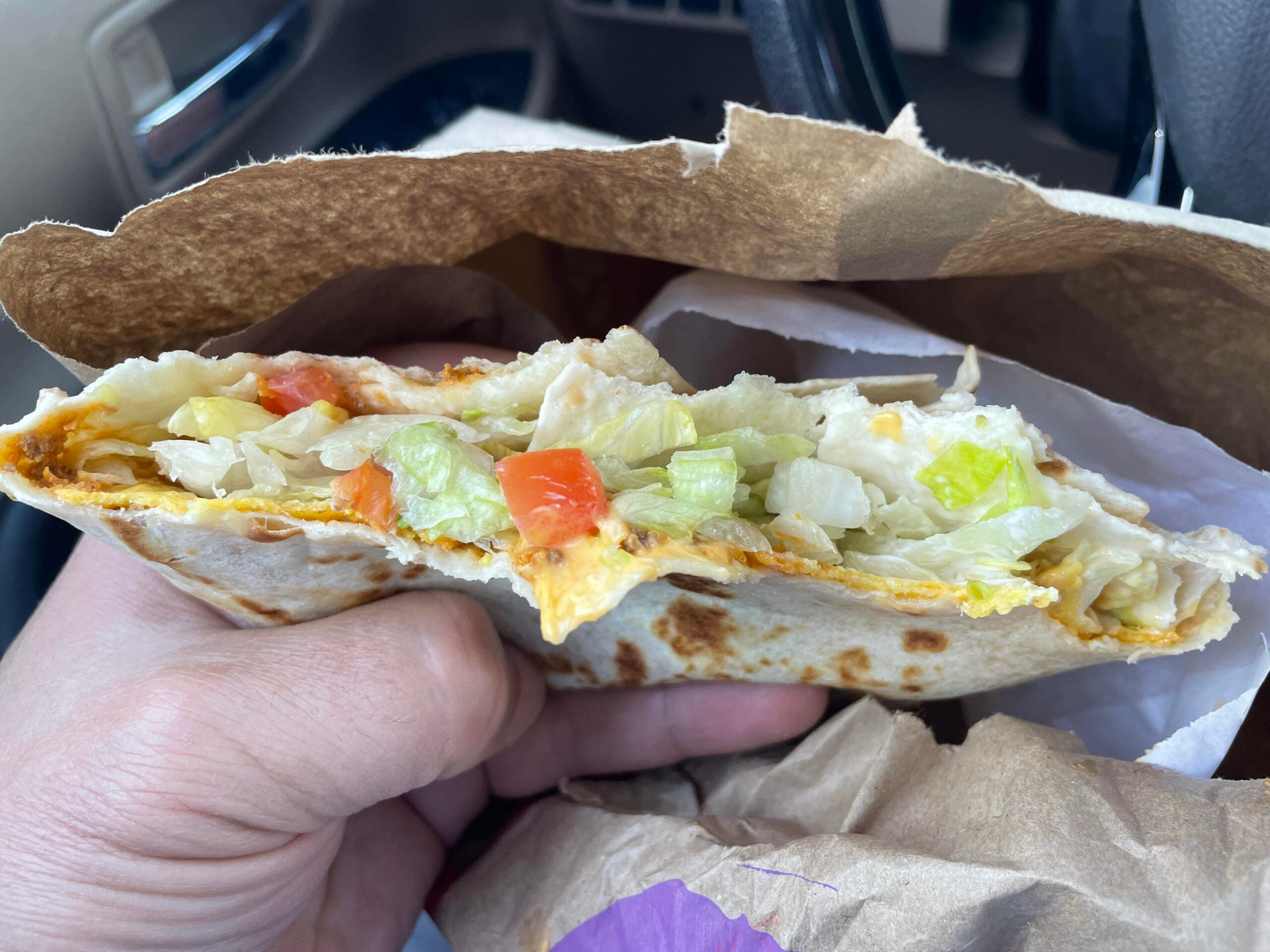 A person holding a folded flour tortilla wrap filled with lettuce, tomatoes, and melted cheese, served in a paper bag.