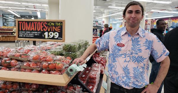 A store employee in a light pink Hawaiian shirt stands next to a display of tomatoes on the vine with a pricing sign above.