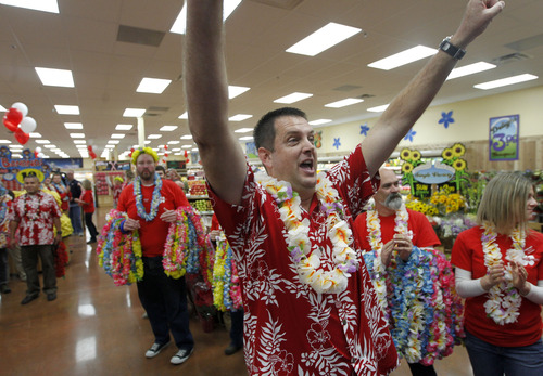 A man in a red floral shirt and flower lei celebrates with his arms raised, surrounded by a group of people wearing matching shirts and leis in a grocery store.