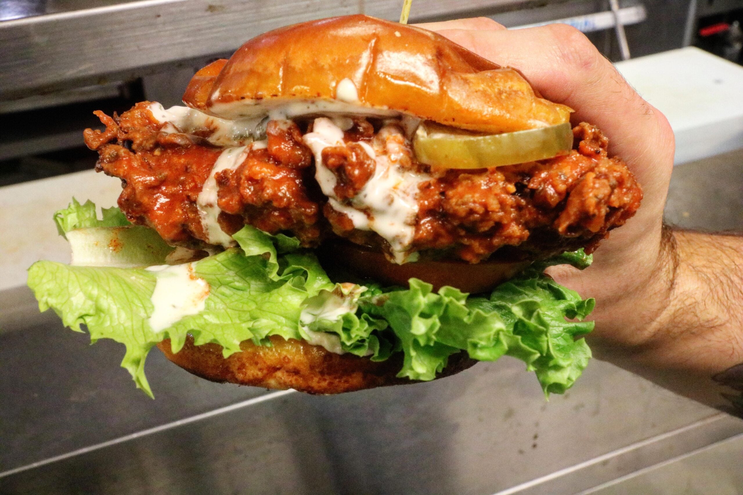 A hand holds a spicy fried chicken sandwich with lettuce, pickles, and drizzled sauce on a bun.