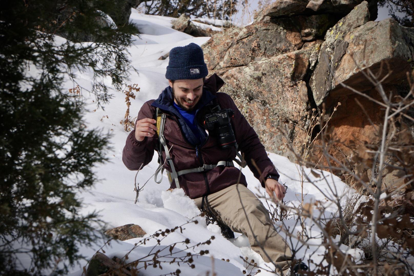A hiker with a camera strapped to his chest smiles while walking through a snowy, rocky landscape.