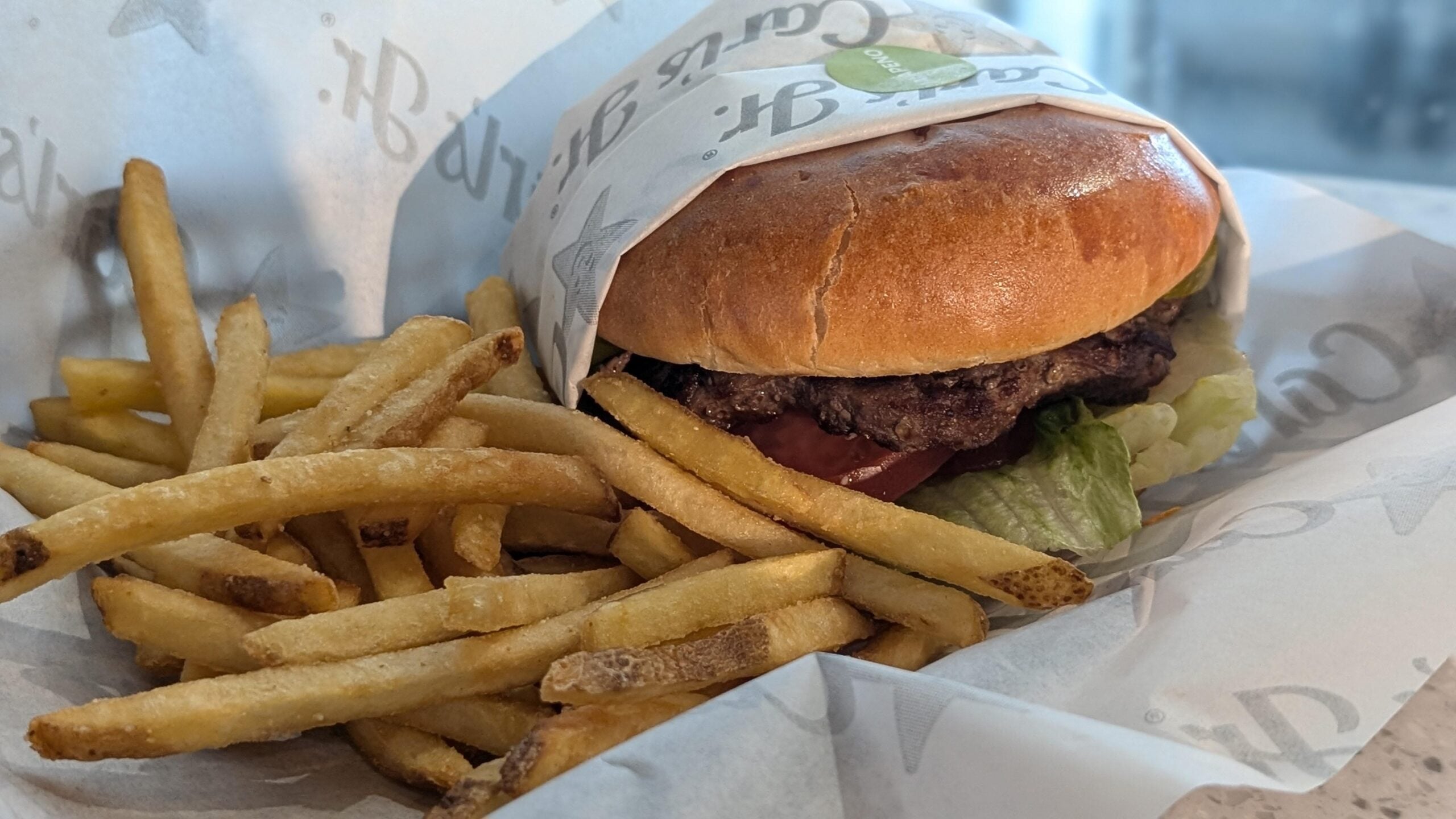 A Carl’s Jr. burger wrapped in branded paper next to a serving of french fries in a paper-lined basket.