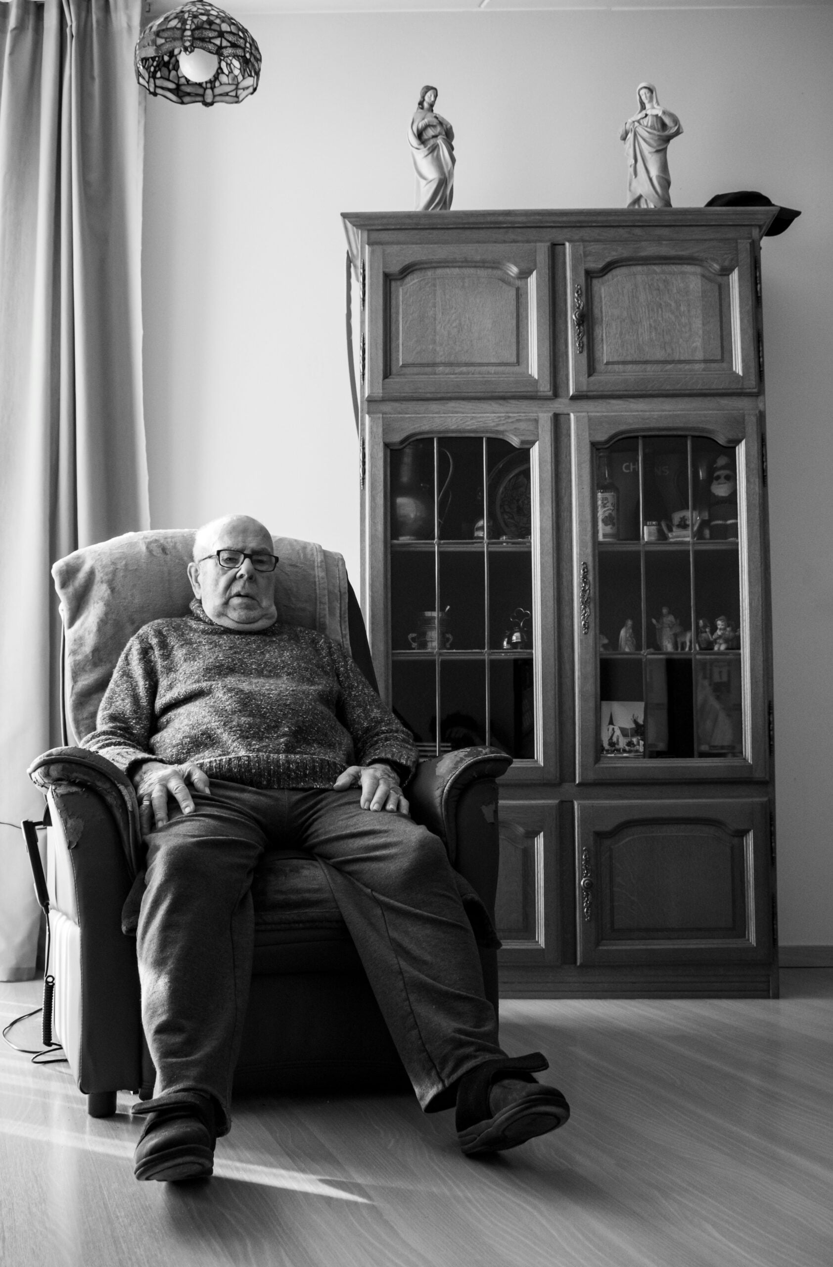An elderly man sits in an armchair in a living room, with a glass-fronted cabinet and two figurines on top behind him. The photo is in black and white.