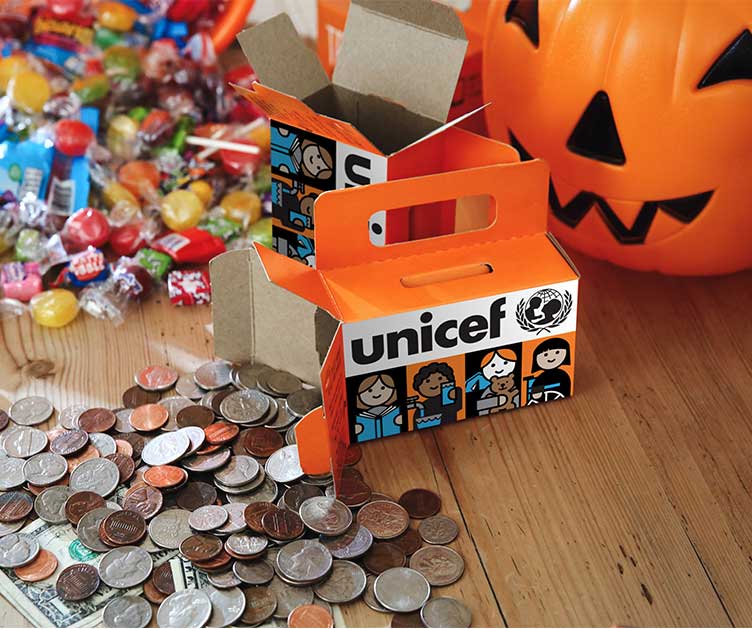 An orange UNICEF cardboard collection box surrounded by a pile of coins, loose candy, and a plastic pumpkin bucket on a wooden table.