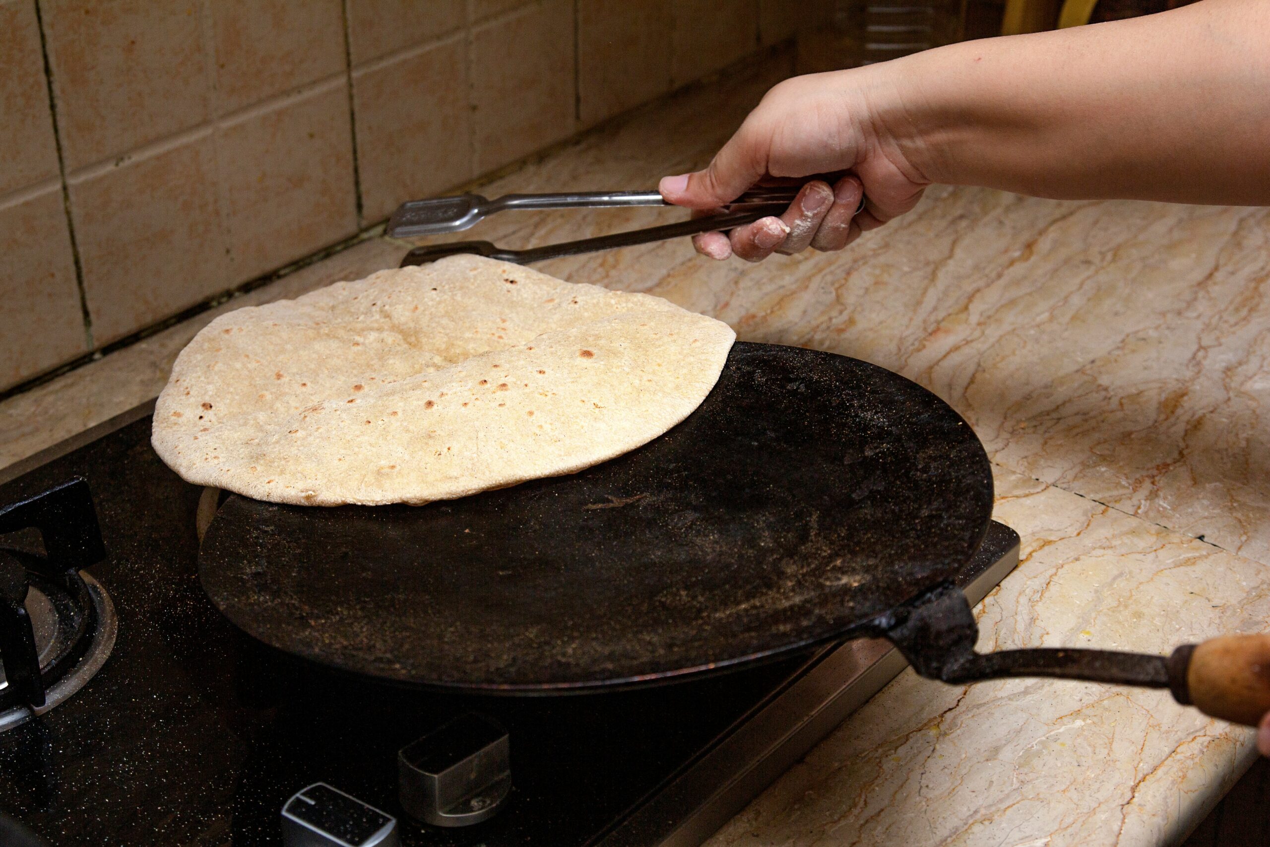 A hand uses tongs to cook a roti on a black iron pan over a gas stove in a kitchen with a beige countertop and tiled wall. Tortillas is one of the affordable foods out there.