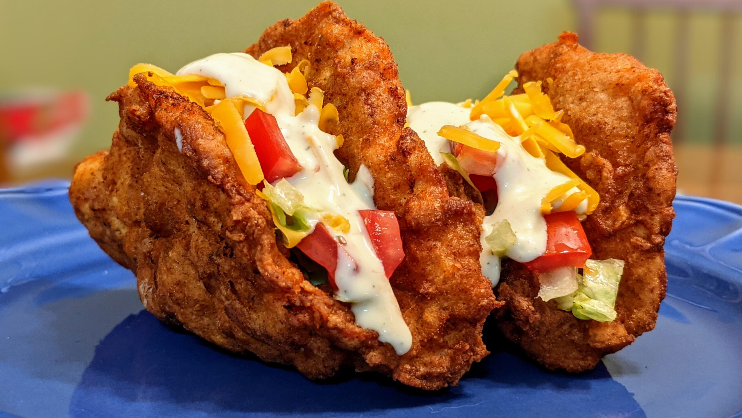 Two tacos with fried chicken shells filled with shredded lettuce, diced tomatoes, shredded cheddar cheese, and drizzled with white dressing.