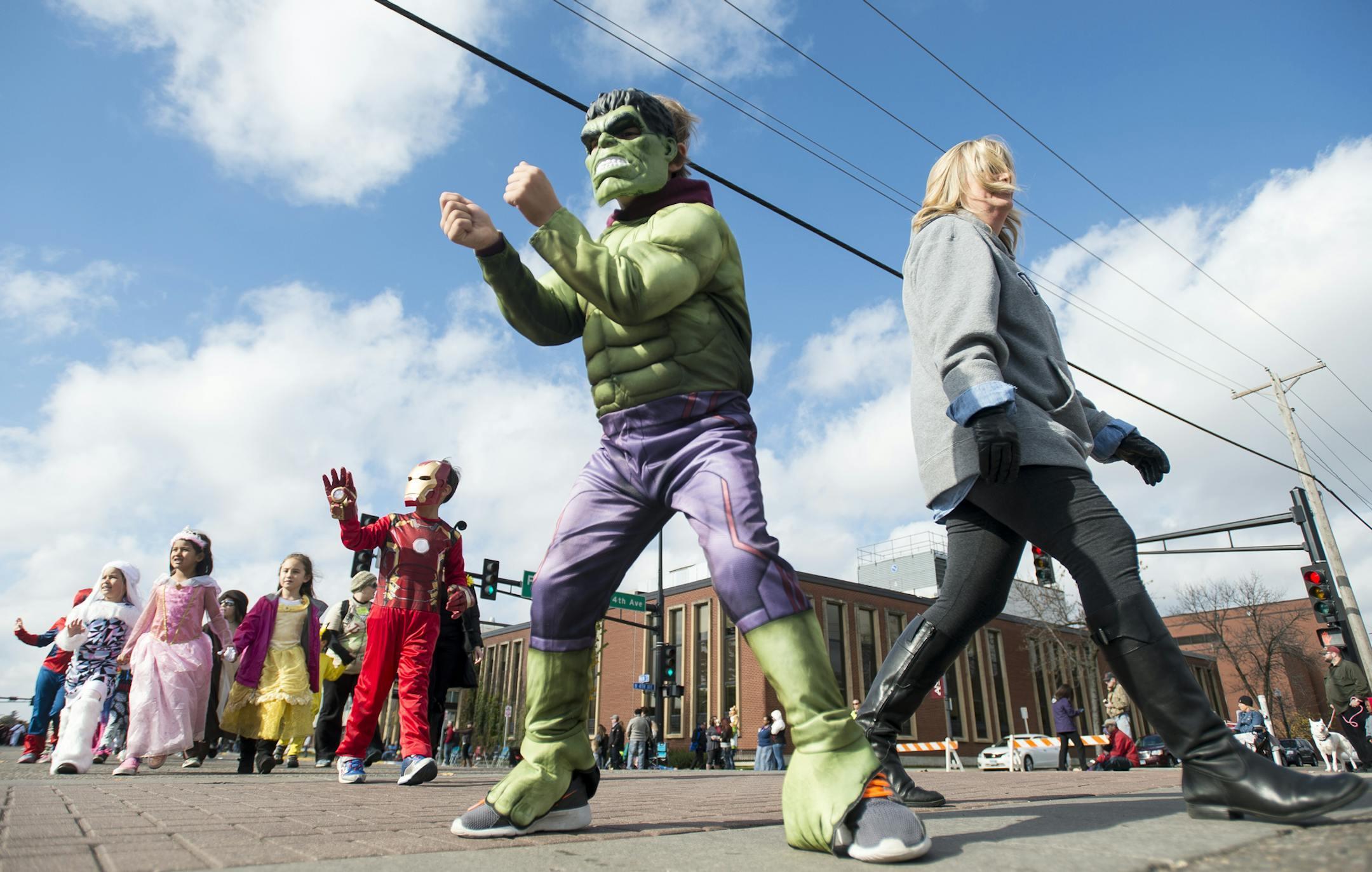 A child in a Hulk costume walks in a parade, followed by a child dressed as Iron Man and other children in costumes, with an adult walking alongside them.