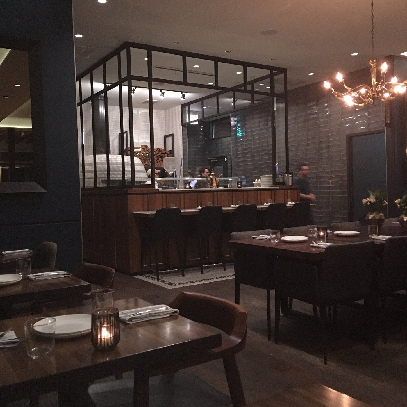A restaurant dining room with dark wooden tables, bar seating, and a glass-enclosed kitchen area with a pizza oven.