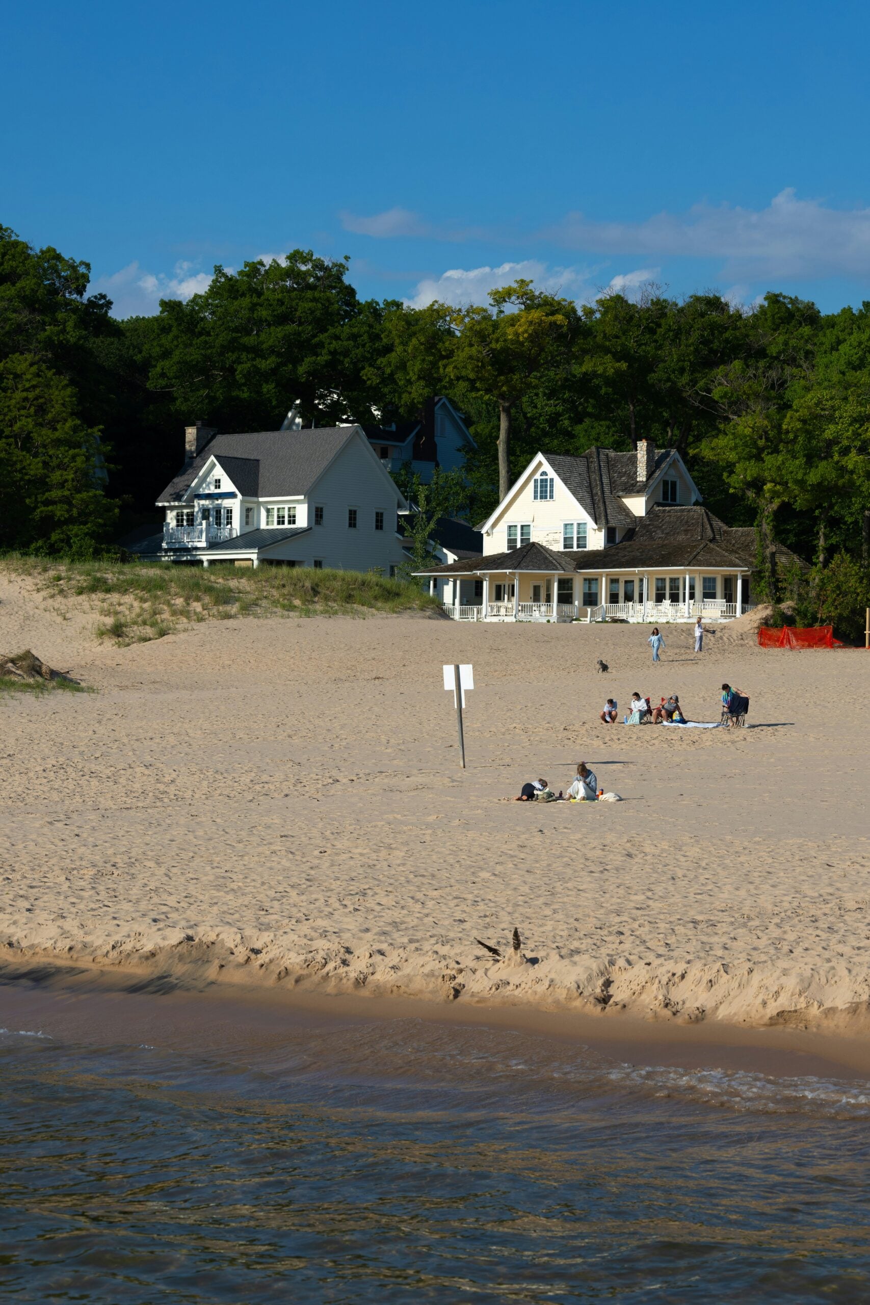 A sandy beach with people sitting and relaxing, two white houses in the background, trees behind the houses, and a signpost in the sand near the shoreline.