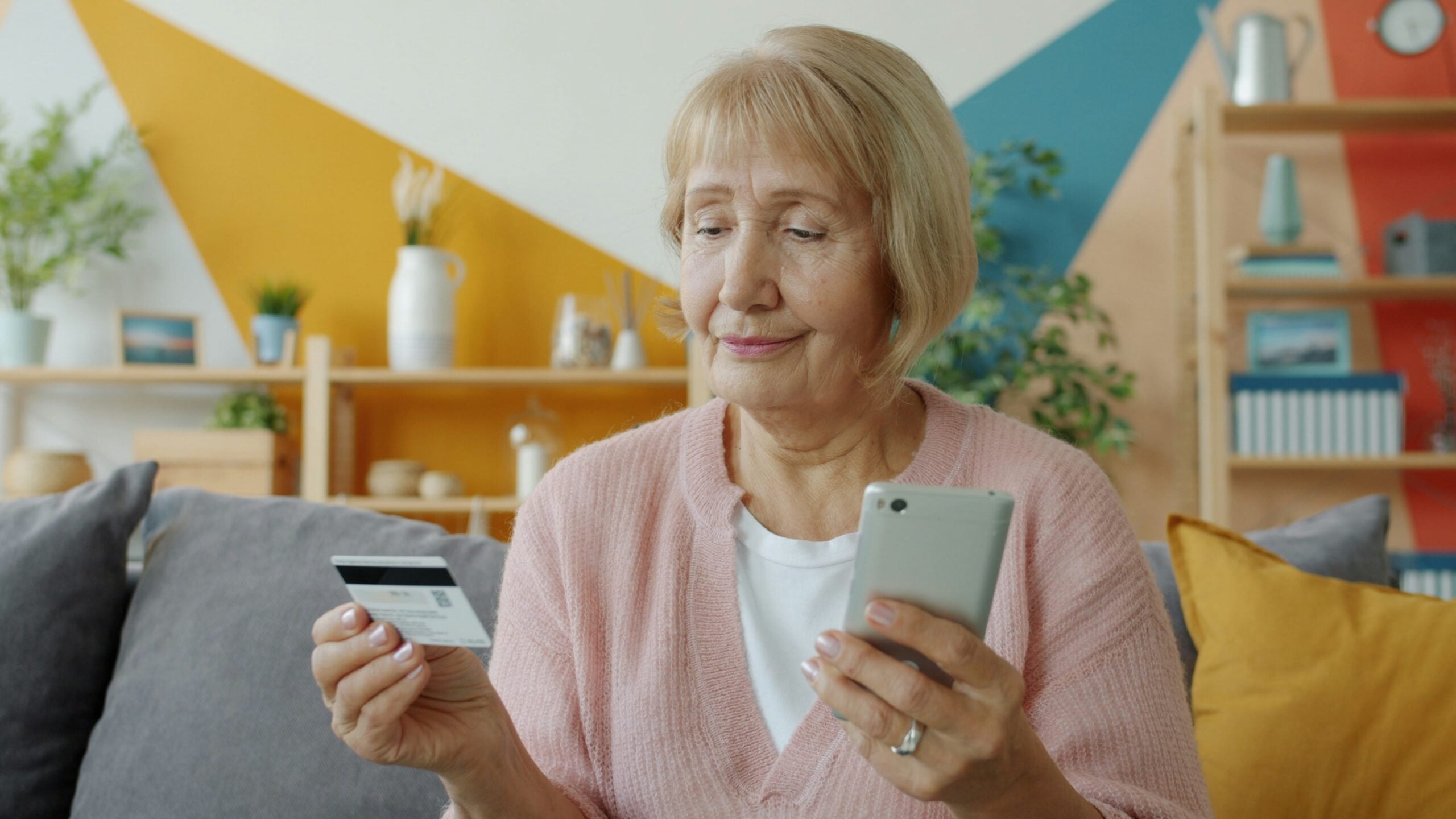 An older woman sits on a couch holding a smartphone in one hand and a credit card in the other, appearing to shop or make a payment online. Shelves and colorful decor are visible in the background.