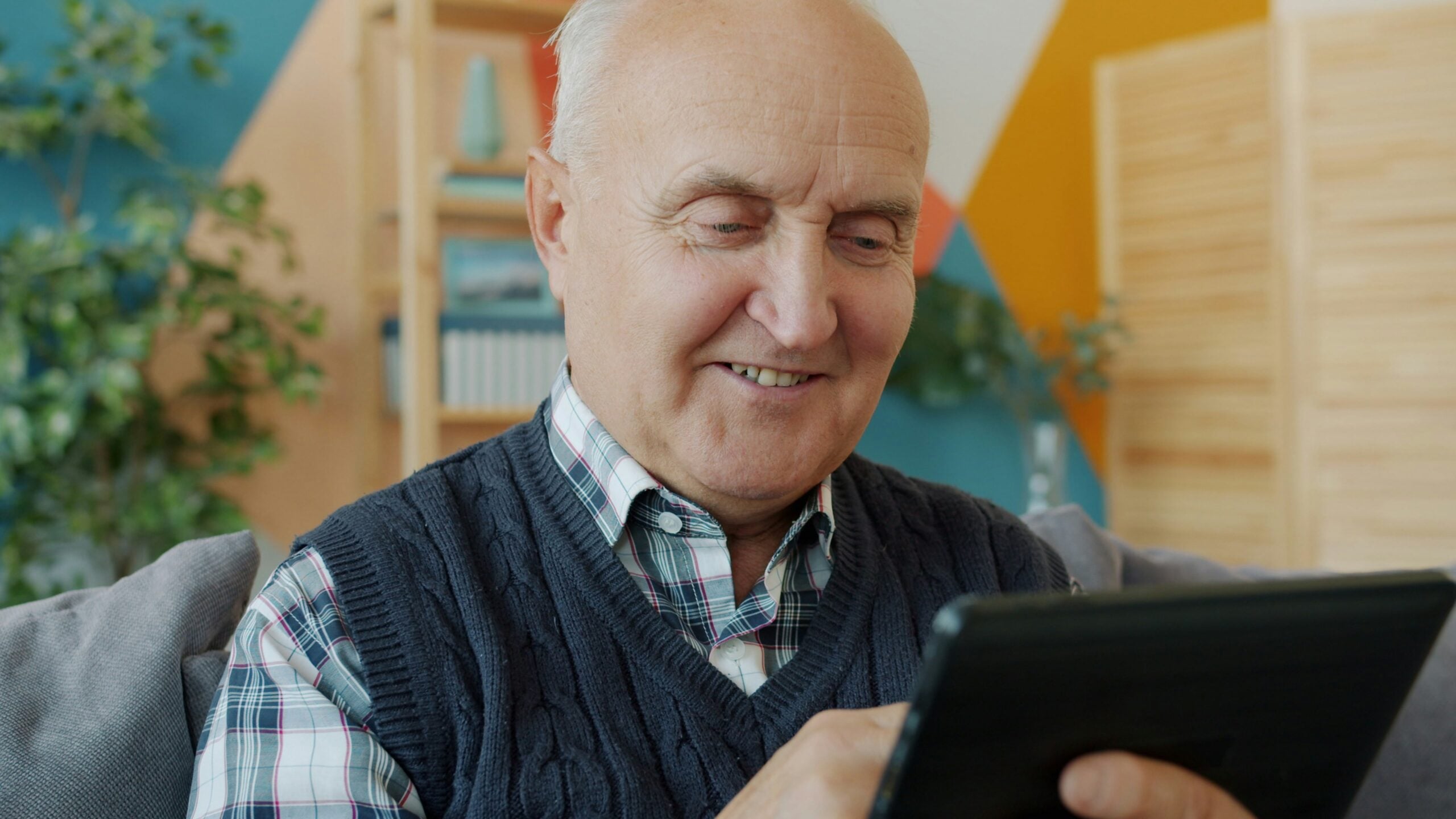 An older man sits on a couch, smiling while using a tablet. He wears a plaid shirt and dark knit vest. Shelves, plants, and colorful walls are visible in the background.