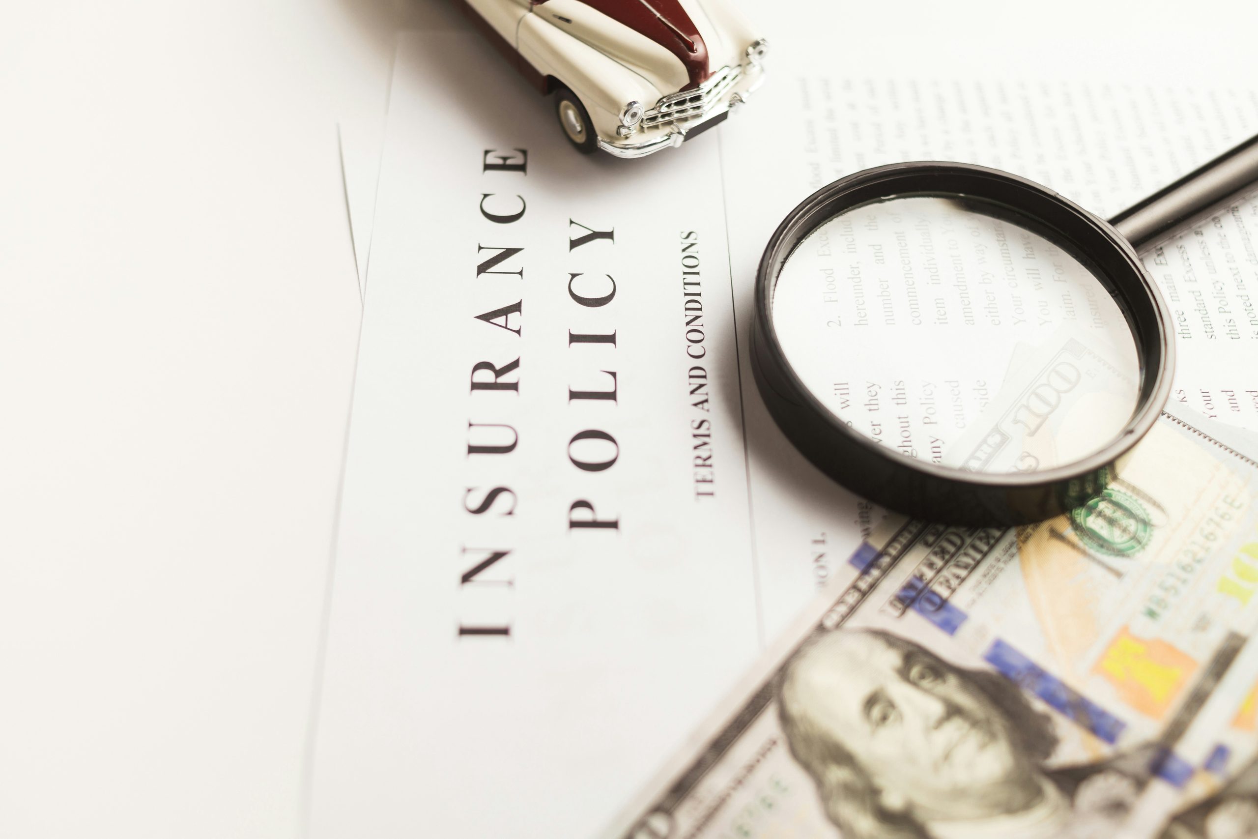 A magnifying glass, a toy car, and a $100 bill rest on an insurance policy document, symbolizing careful review of insurance terms and financial considerations.