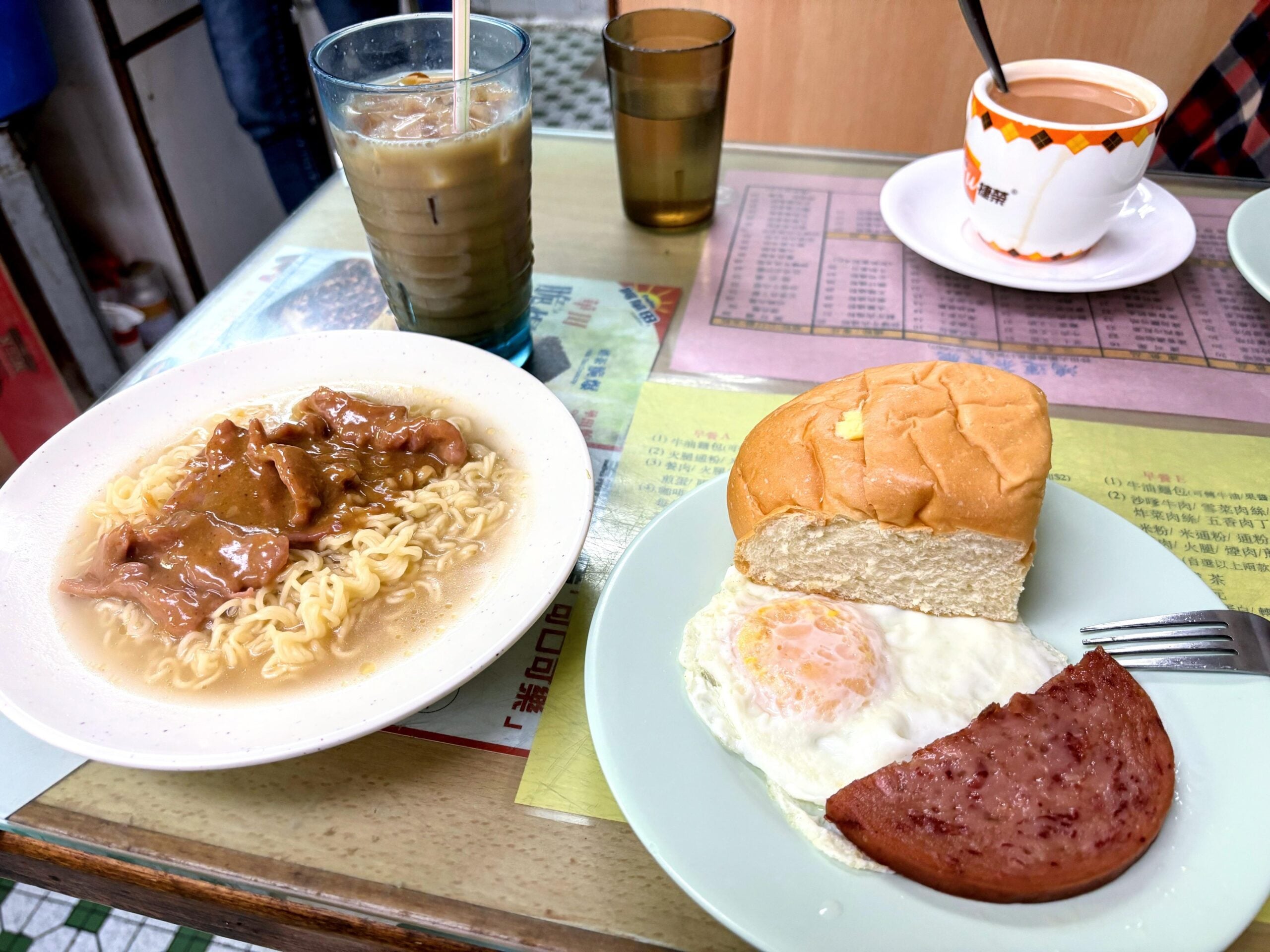 A plate of instant noodles with meat, a plate with a bun, fried egg, and a slice of luncheon meat, and an iced coffee on a restaurant table.