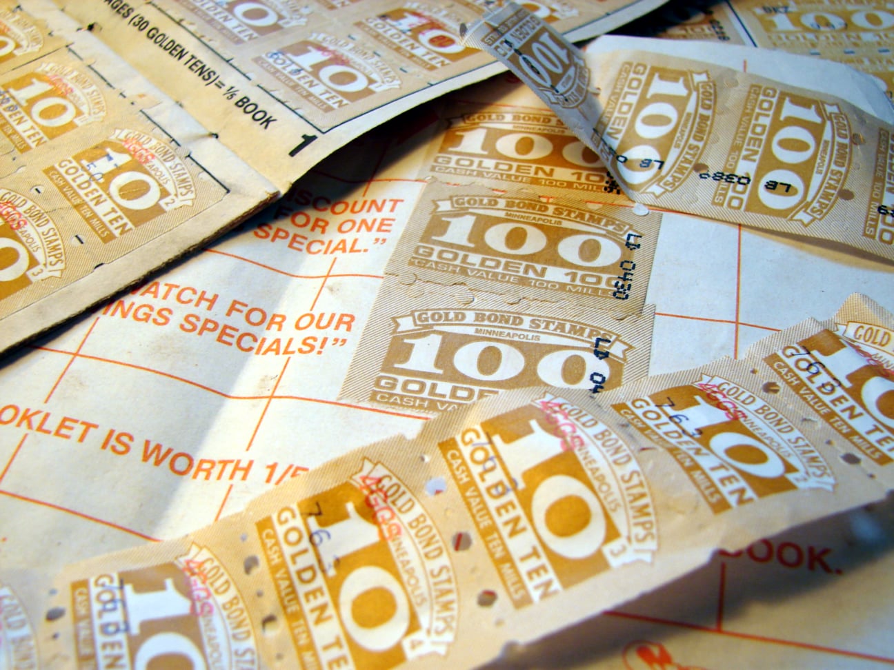 Sheets and strips of vintage Gold Bond trading stamps with the text “10” and “100” printed on a paper background.
