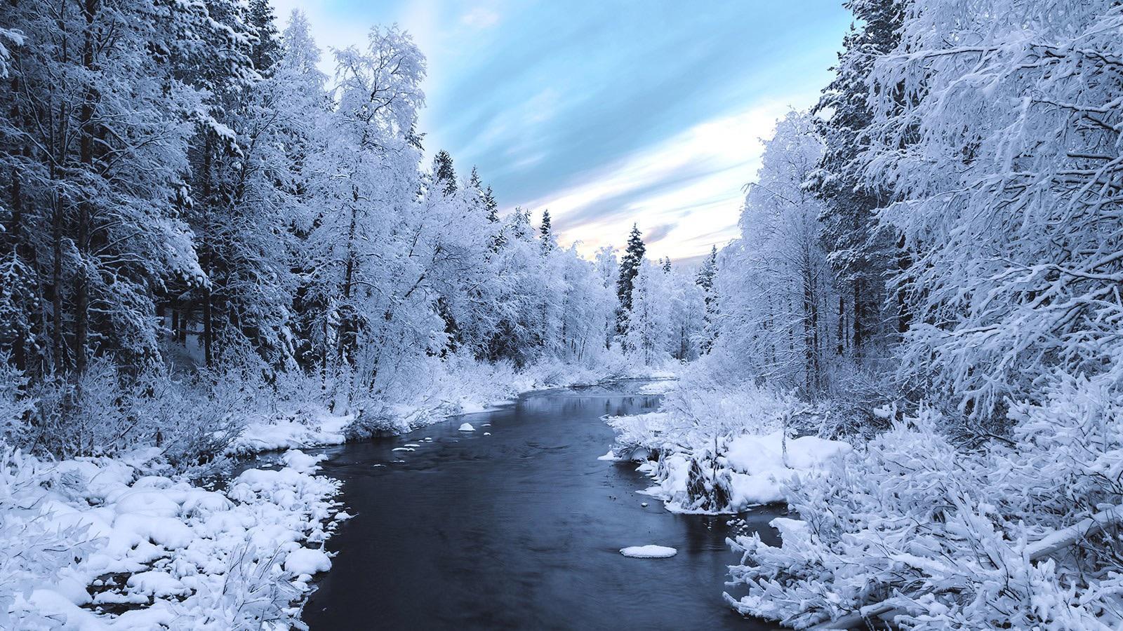 A narrow, dark river flows through a snow-covered forest with frost-laden trees under a pale sky.