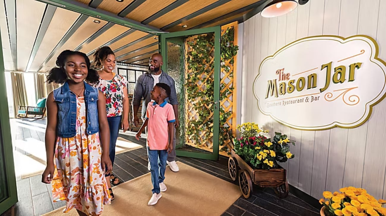 A family walks into The Mason Jar restaurant entrance, marked by a large wall sign and a wooden cart filled with yellow flowers.