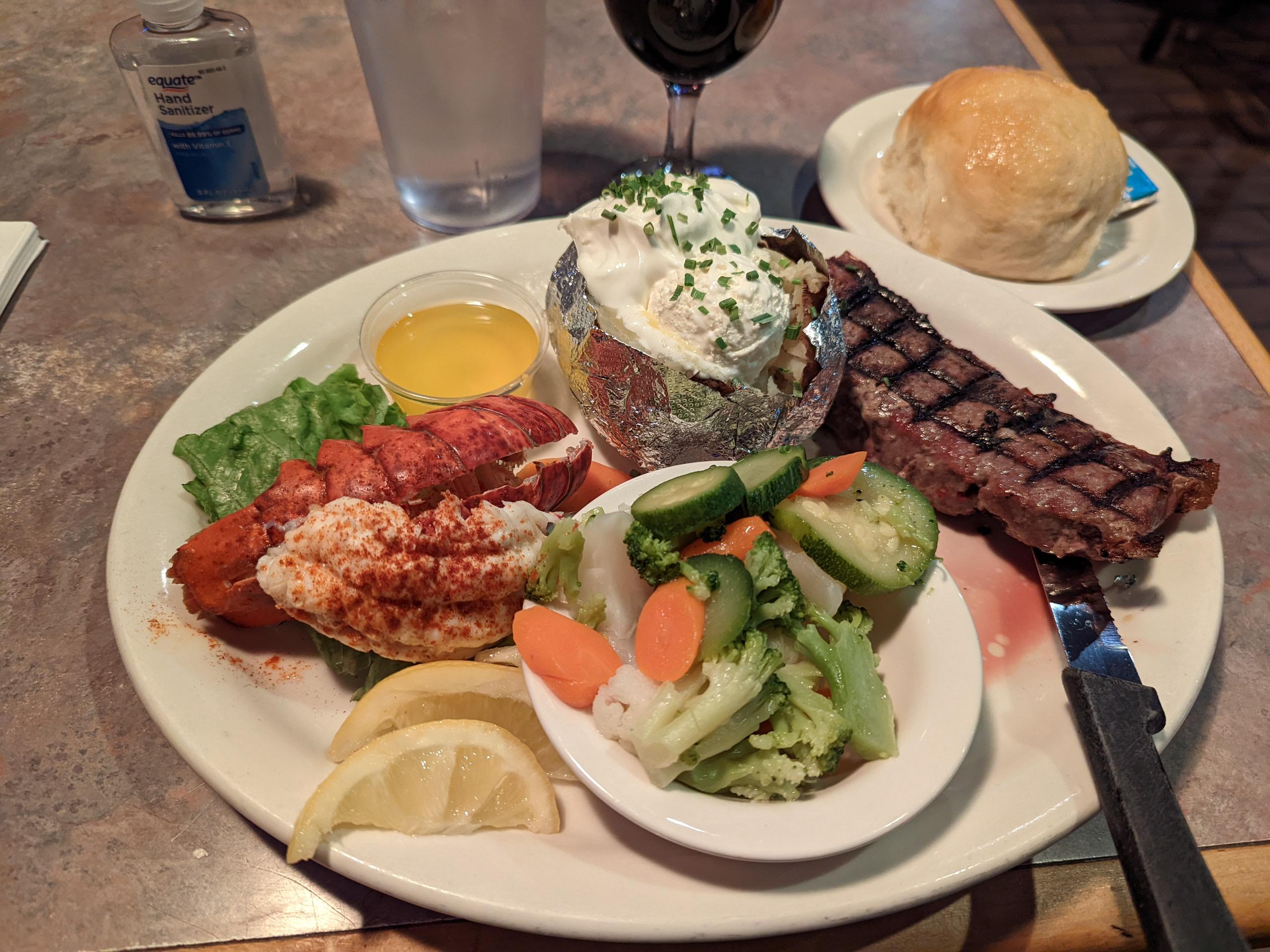Surf and turf dinner plate featuring a grilled steak, lobster tail with butter, a baked potato with sour cream, and a side of steamed vegetables, served with a dinner roll.