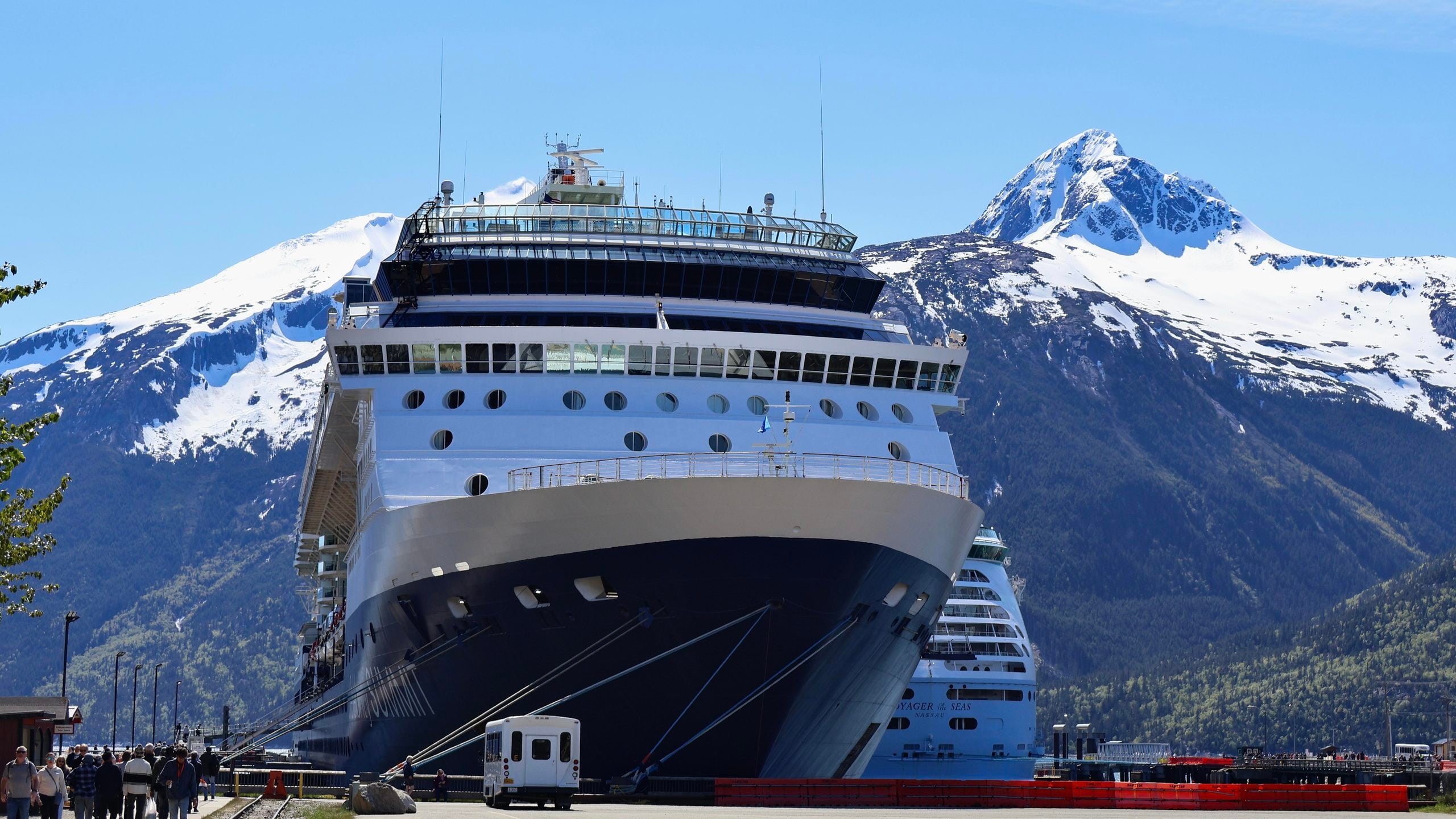 A large cruise ship docked at a port with snow-capped mountains in the background.