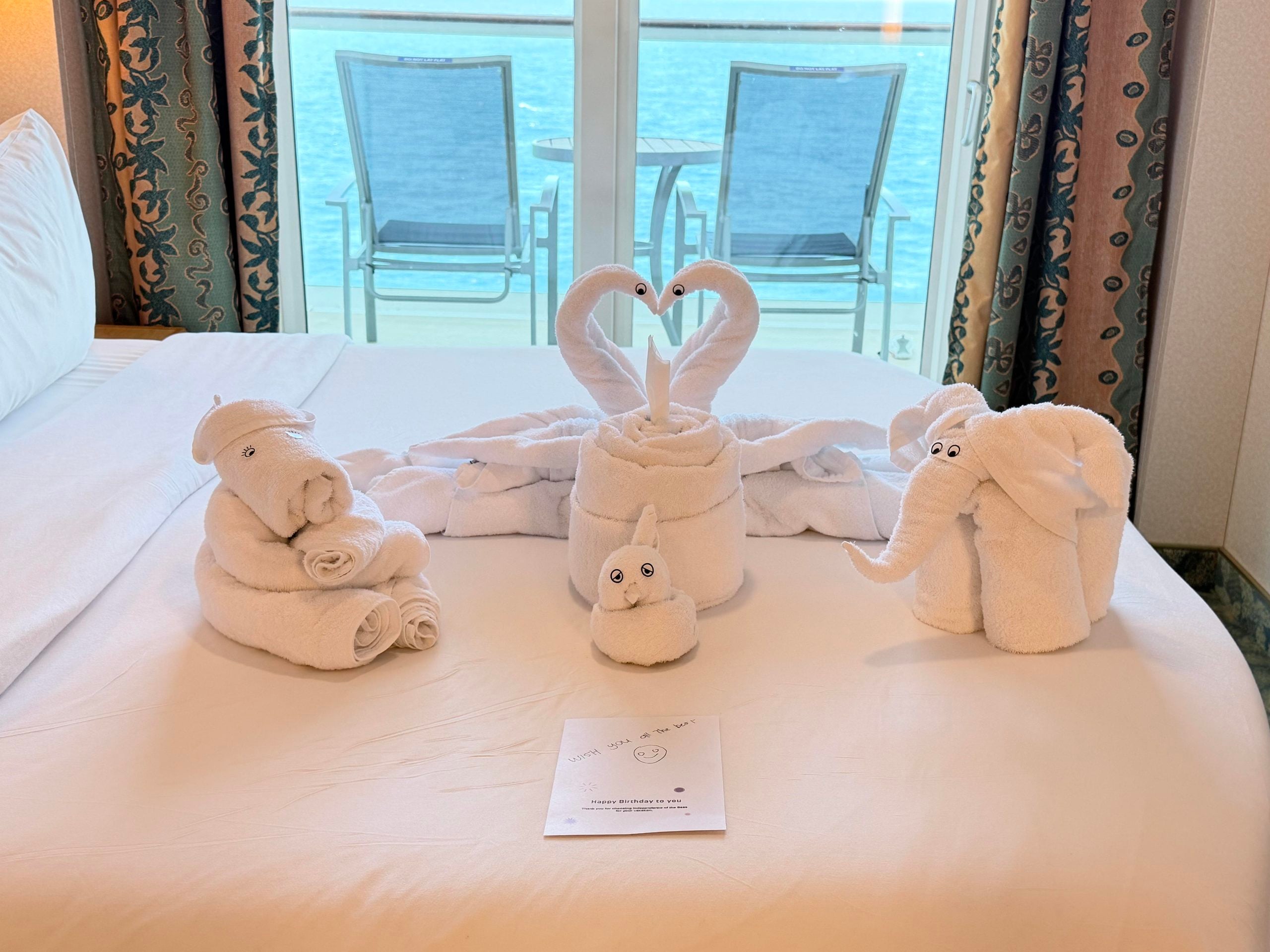 Towel animals shaped like two swans, an elephant, and a small bird displayed on a cruise ship bed in front of a balcony overlooking the ocean, with a birthday card placed in the center.