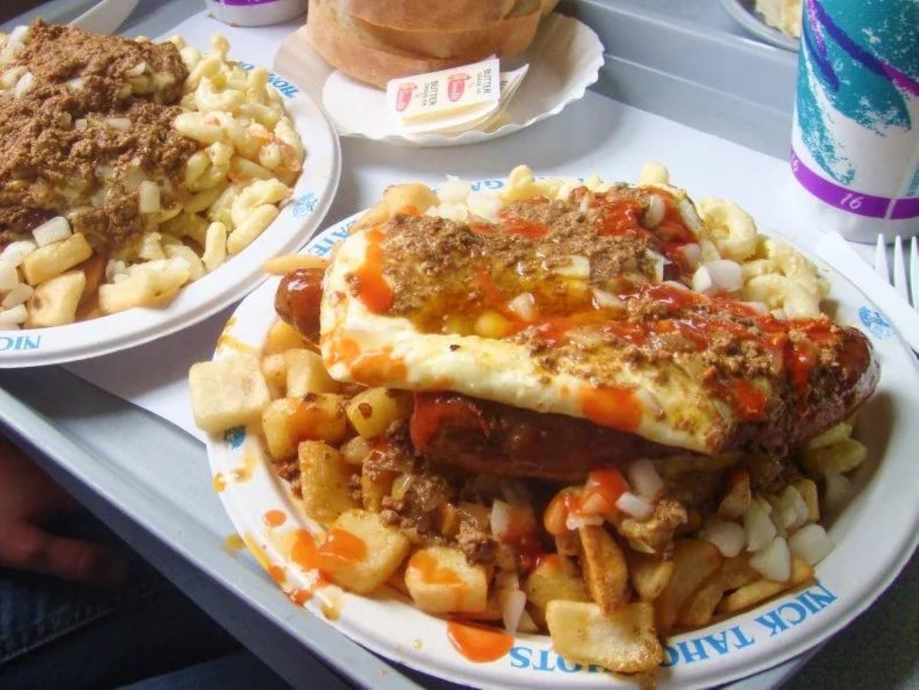 Two plates of “garbage plates” featuring hot dogs, macaroni salad, and home fries, topped with meat sauce, diced onions, and hot sauce.