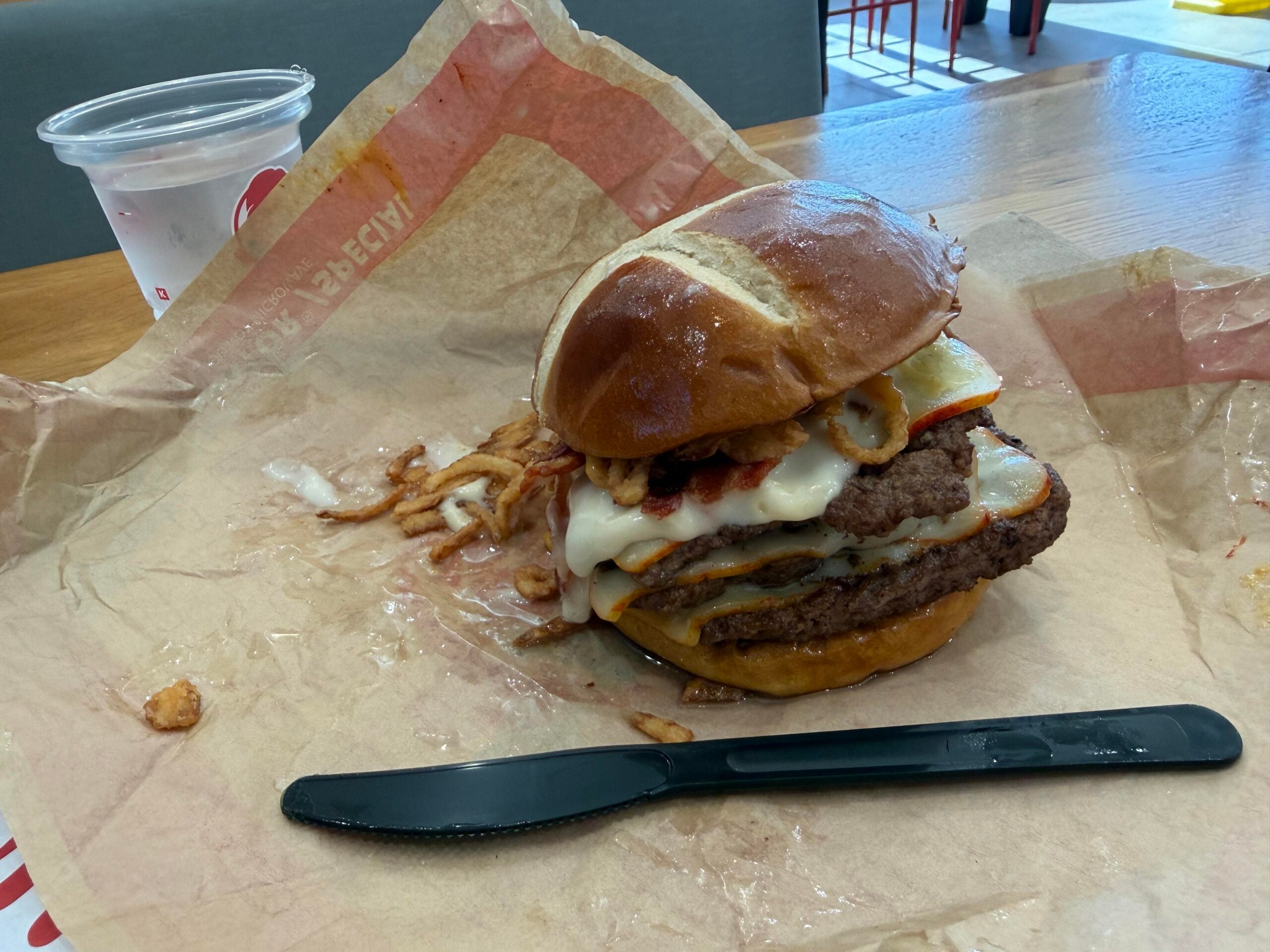 A double cheeseburger with bacon and crispy onions on a pretzel bun, served on branded parchment paper with a plastic knife and a cup of water.