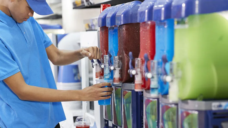 A person in a blue polo shirt fills a plastic cup from a slushy machine with multiple colorful frozen drink dispensers.