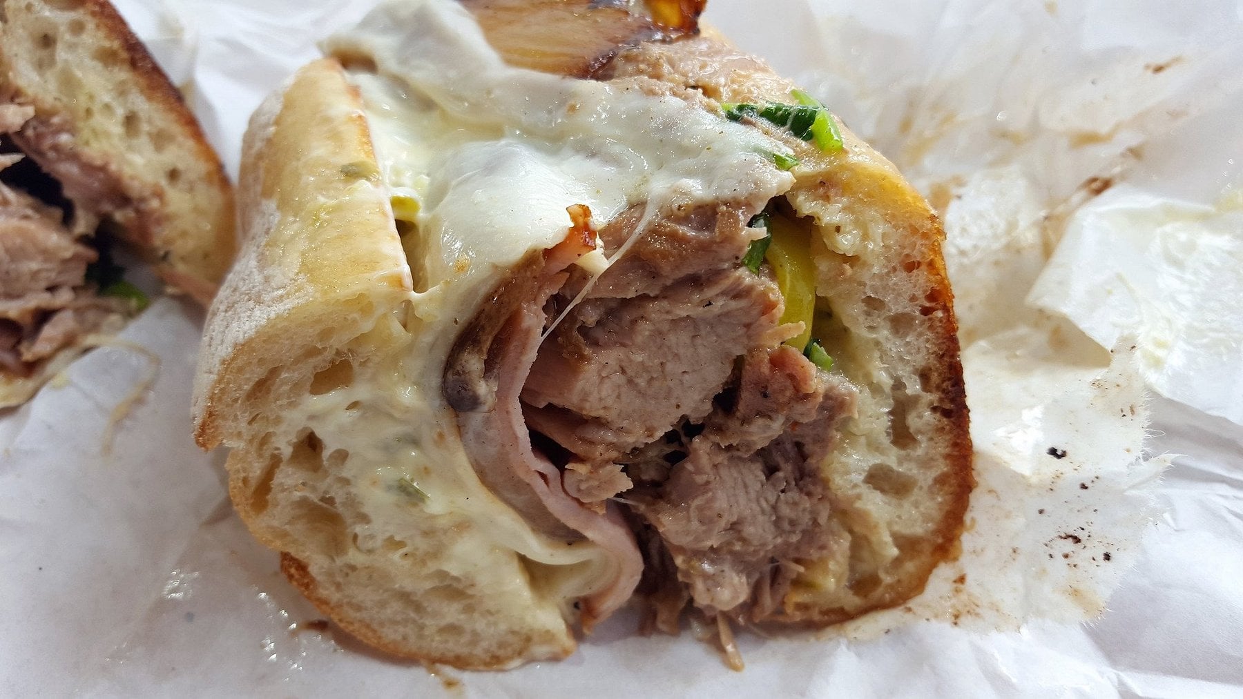 A close-up of a partially eaten Cuban sandwich on parchment paper, featuring roasted pork, ham, melted cheese, and pickles on toasted bread.