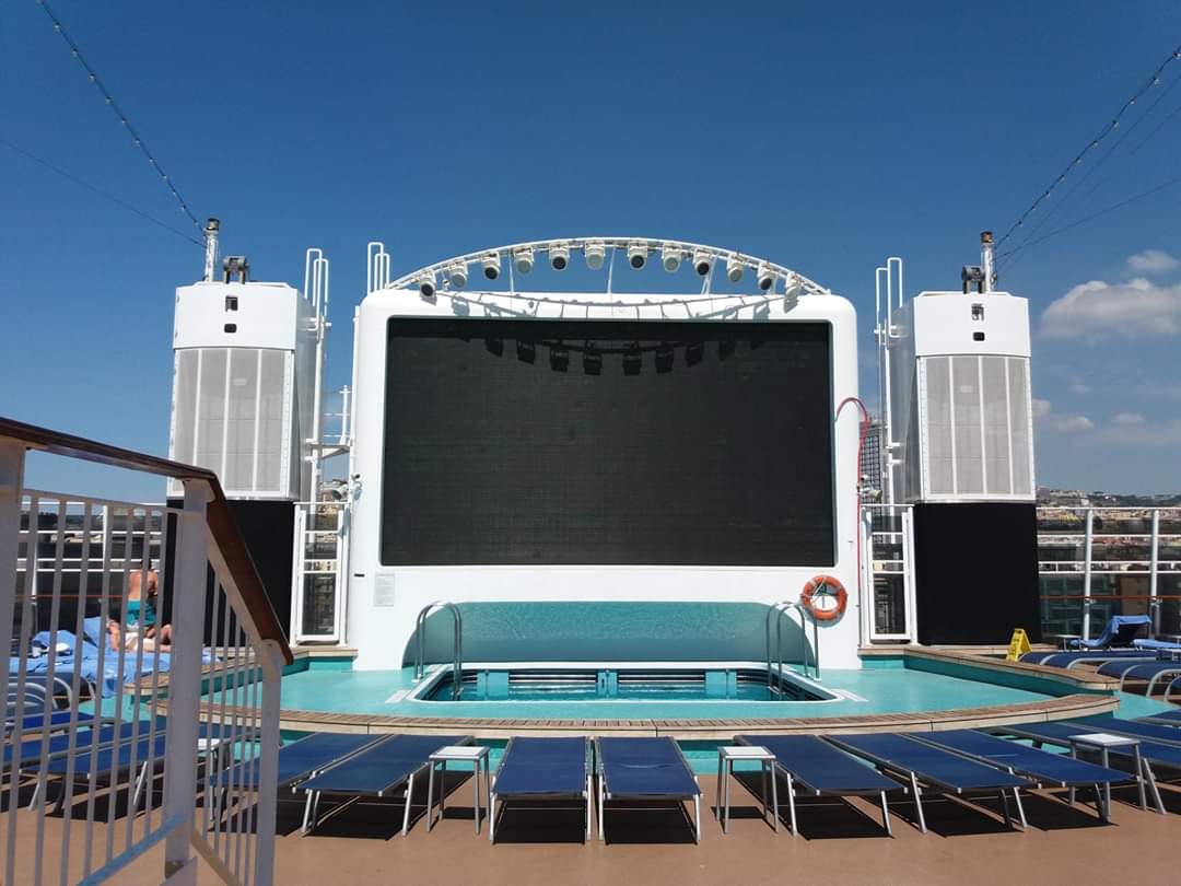 A large outdoor movie screen and hot tub on a cruise ship deck surrounded by lounge chairs.