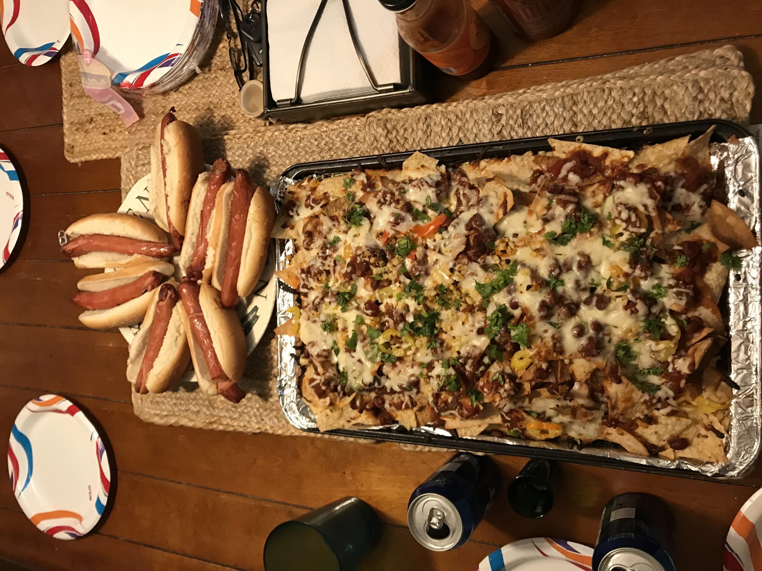 A tray of loaded nachos with melted cheese and greens beside a plate of hot dogs on buns, paper plates, soda cans, and condiments on a wooden table.