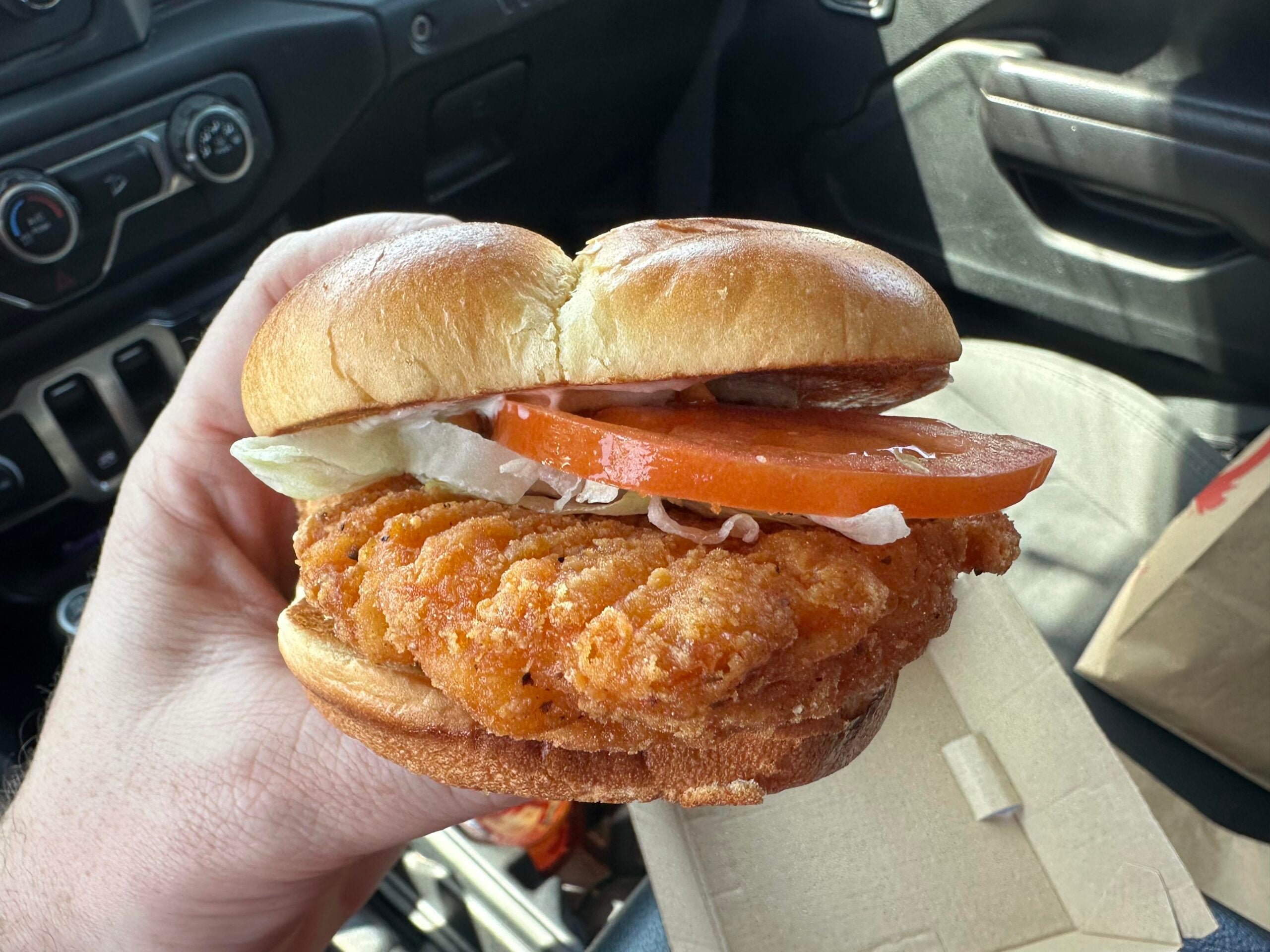 A fried chicken sandwich with lettuce and a tomato slice, held by hand inside a vehicle.