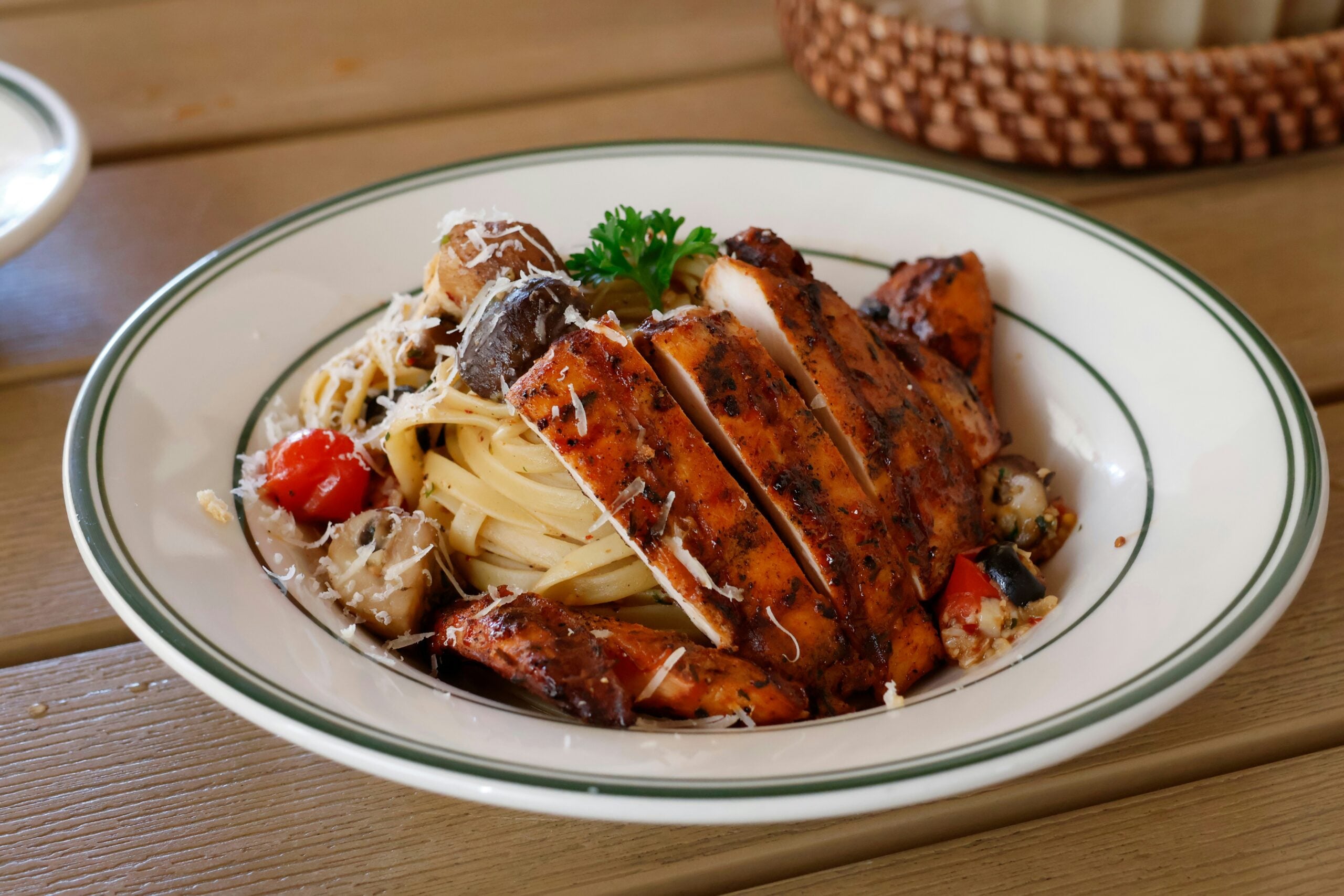 A bowl of linguine pasta topped with sliced grilled chicken, mushrooms, cherry tomatoes, and shaved cheese.