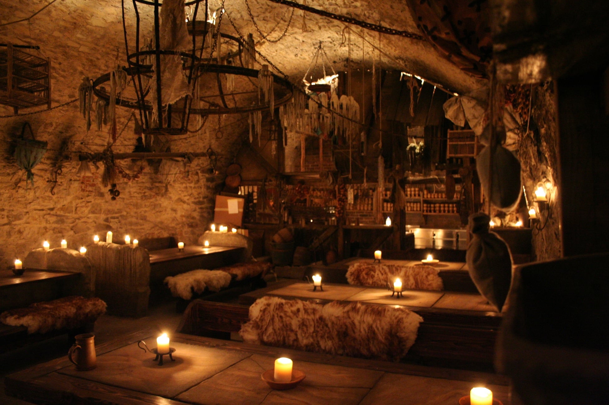 A dimly lit medieval tavern with stone walls, wooden tables, benches covered in furs, candles, and rustic decor creates a warm and cozy atmosphere.