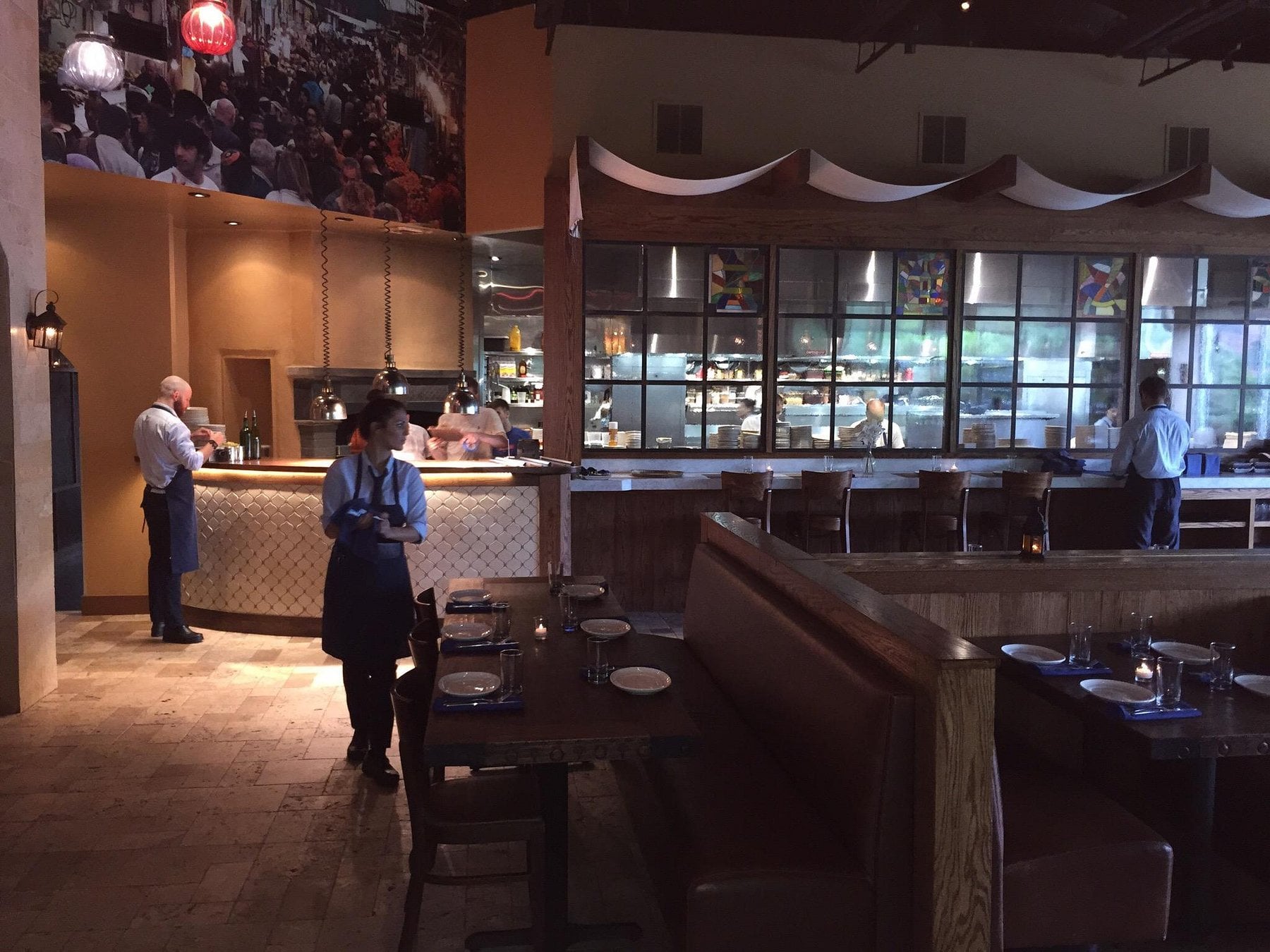 A server in a dark apron stands before dining tables and bar seating in a restaurant, with a kitchen visible through large glass windows in the back.