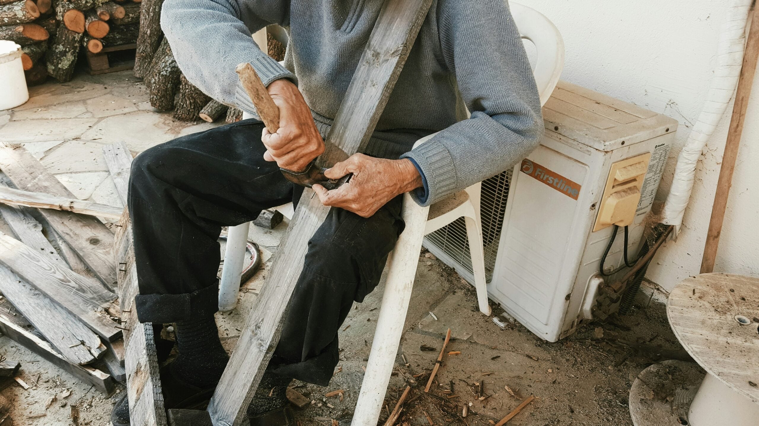An older person sits on a plastic chair outdoors, carving a piece of wood with a hand tool. Wood shavings and planks are scattered on the ground around them. Their face is not visible.