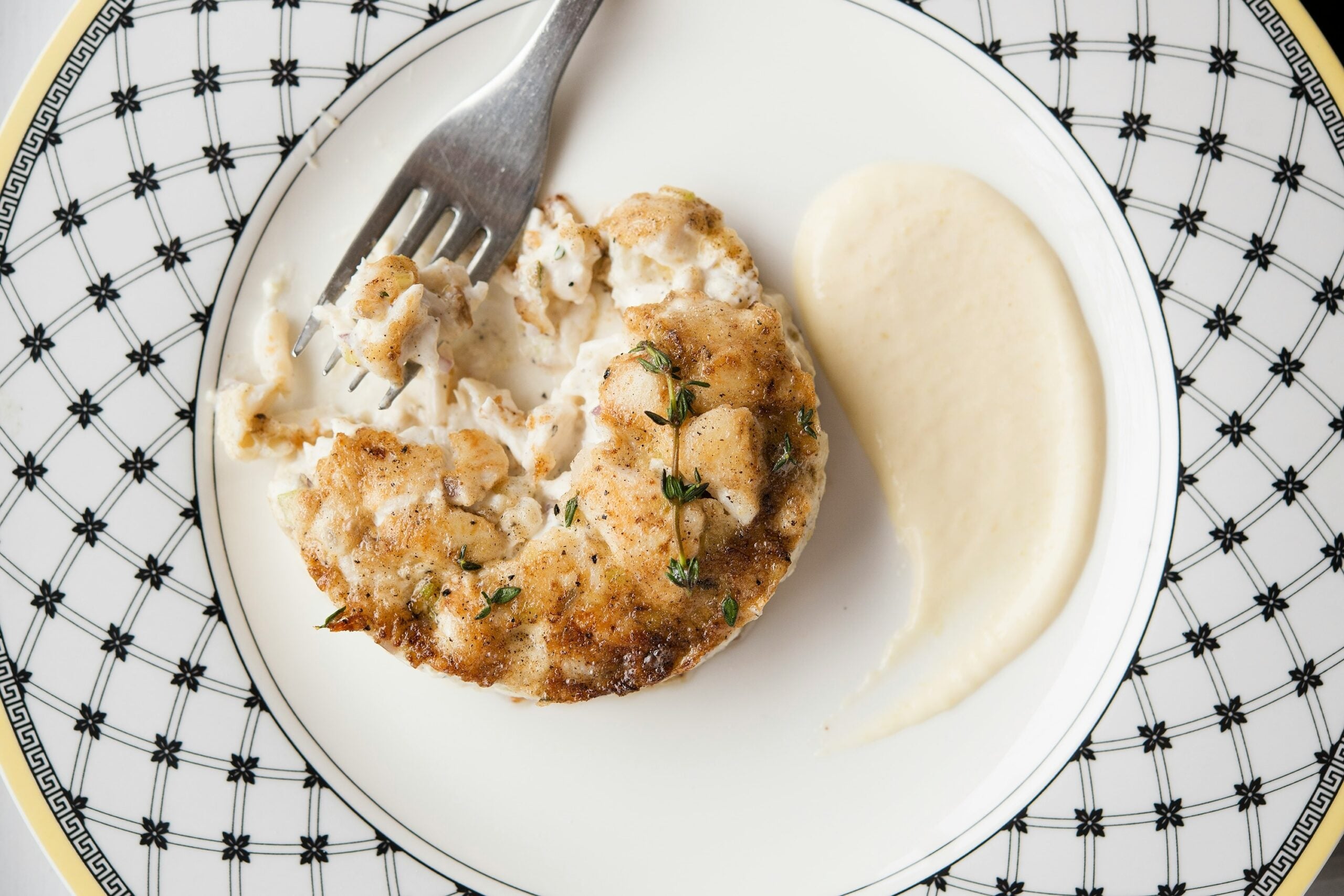 A crab cake topped with fresh herbs on a patterned plate, served with a side of creamy sauce and a fork.