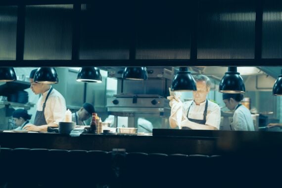 Chefs in white uniforms and aprons work in a restaurant kitchen behind a counter under hanging pendant lights, preparing restaurant-made dishes.