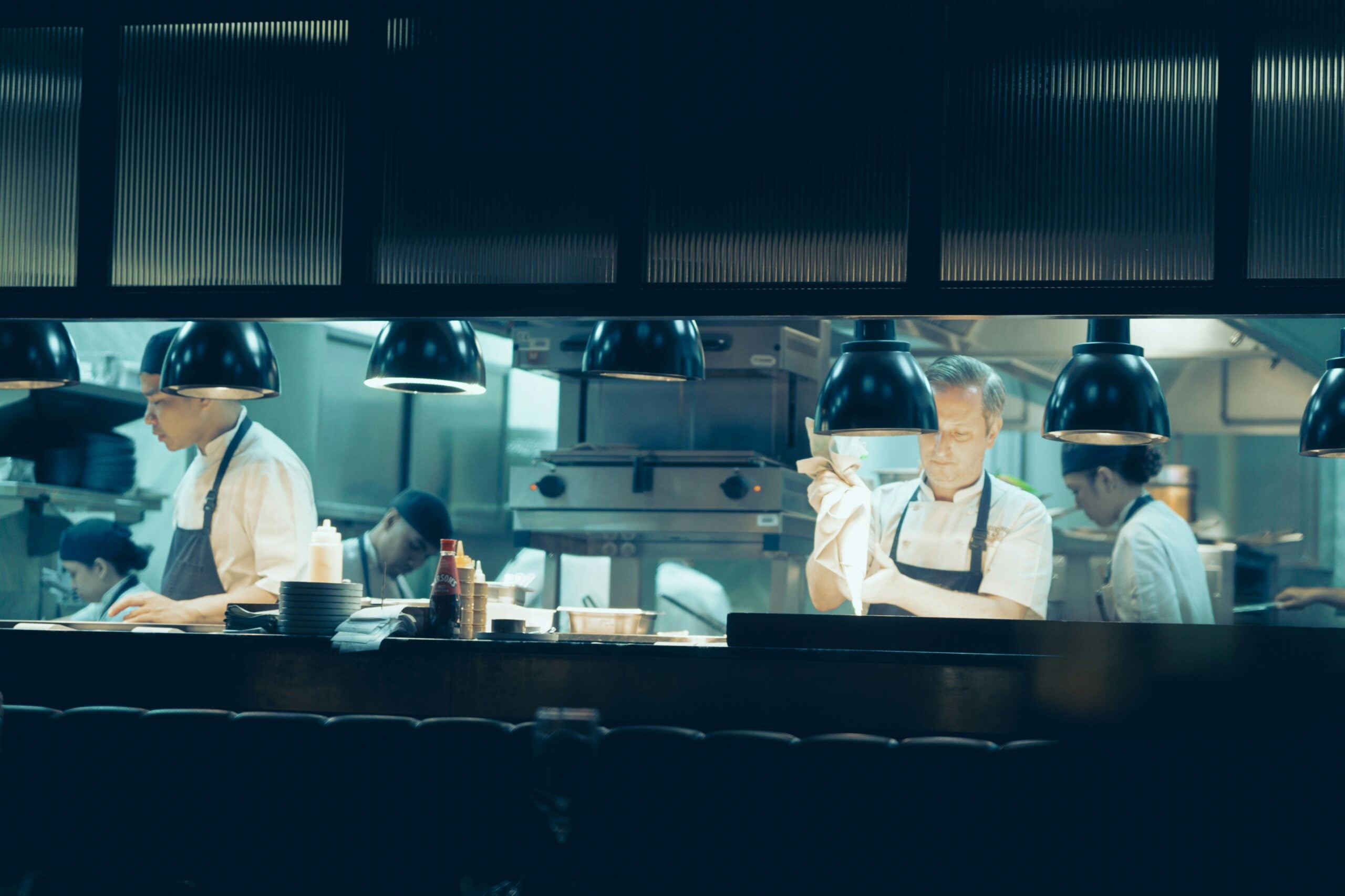 Chefs in white uniforms and aprons work in a restaurant kitchen behind a counter under hanging pendant lights, preparing restaurant-made dishes.