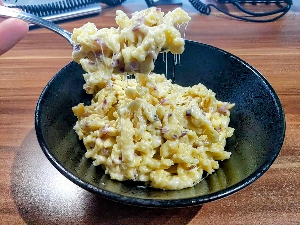 A metal fork lifts a portion of cheesy spaetzle with red onion from a black bowl, with strands of melted cheese stretching between them.